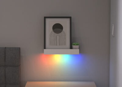 A shelf on a wall holding a framed art print and plant, with a rainbow colored light shining from the bottom.
